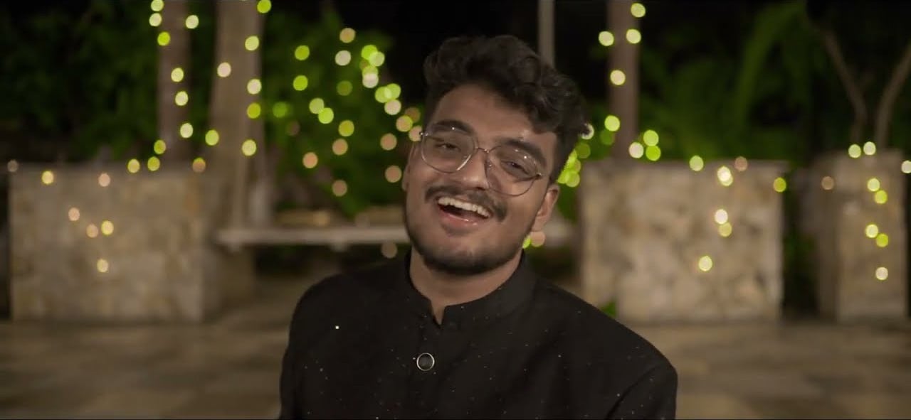 JP Kalaravkar in a black bandhgala, smiling as he sings among fairy lights in the Mare Palavde Bandhayo video