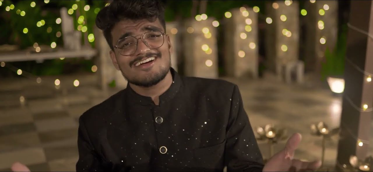 JP Kalaravkar singing with an open hand in the Mare Palavde Bandhayo video, lamps and lights behind him